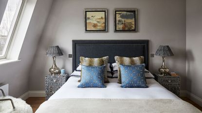 A double attic bedroom space with taupe walls, a double bed with dark navy headboard, and white bedding with assorted cushions. There are matching nightstands and lamps to either side of the bed.