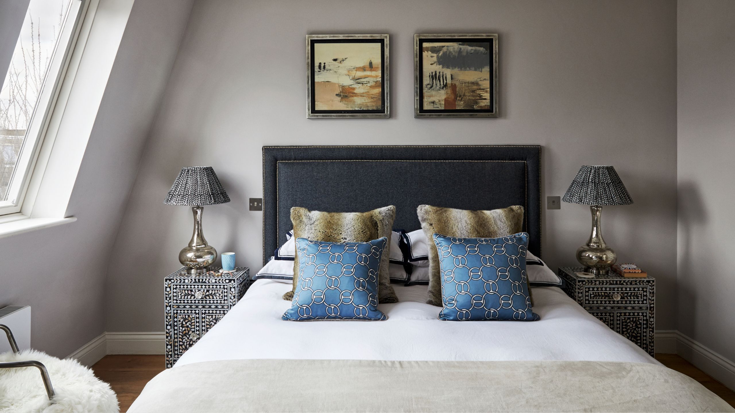 A double attic bedroom space with taupe walls, a double bed with dark navy headboard, and white bedding with assorted cushions. There are matching nightstands and lamps to either side of the bed.