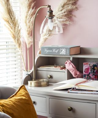 A light gray home office desk with books on top of it and a brass table lamp on top of it, a pink wall behind it with three brown pampas grass stems, and a gray armchair with a mustard yellow throw pillow in front