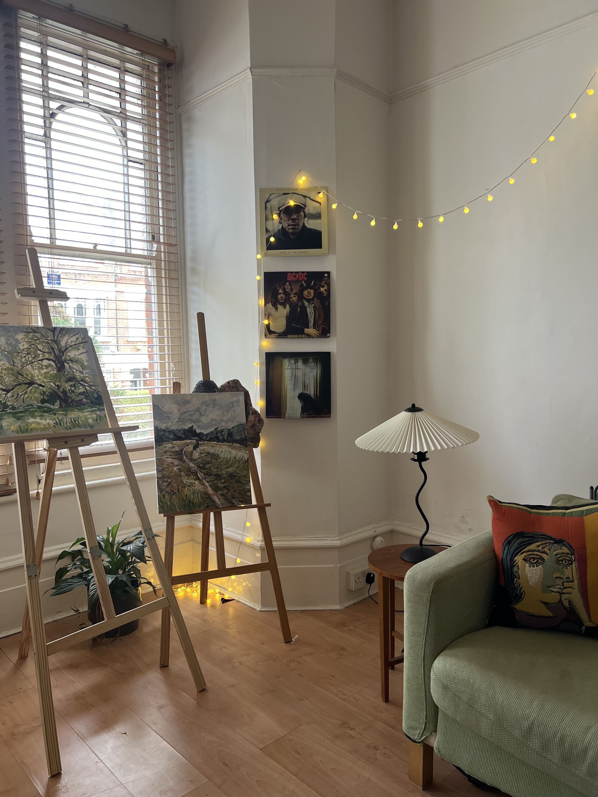 Image of the corner of a living room with wooden floors, high ceilings, and white walls that have crown molding. There are three vinyls styled on the wall on acrylic shelving. There are two easels with paintings on them, the corner of a sage green sofa in view and a wooden side table with a wavy lamp on it.
