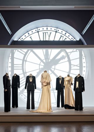 View of Musée d'Orsay's tribute to Yves Saint Laurent featuring seven mannequins displaying a mixture of black suits and beige dresses on a platform in a space with wood flooring and a large clock
