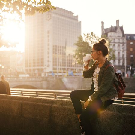 Woman drinking coffee while sitting retailing wall of bridge in city
