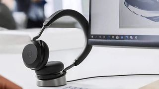 A Jabra headset sitting in its charger dock on a desk next to a PC monitor