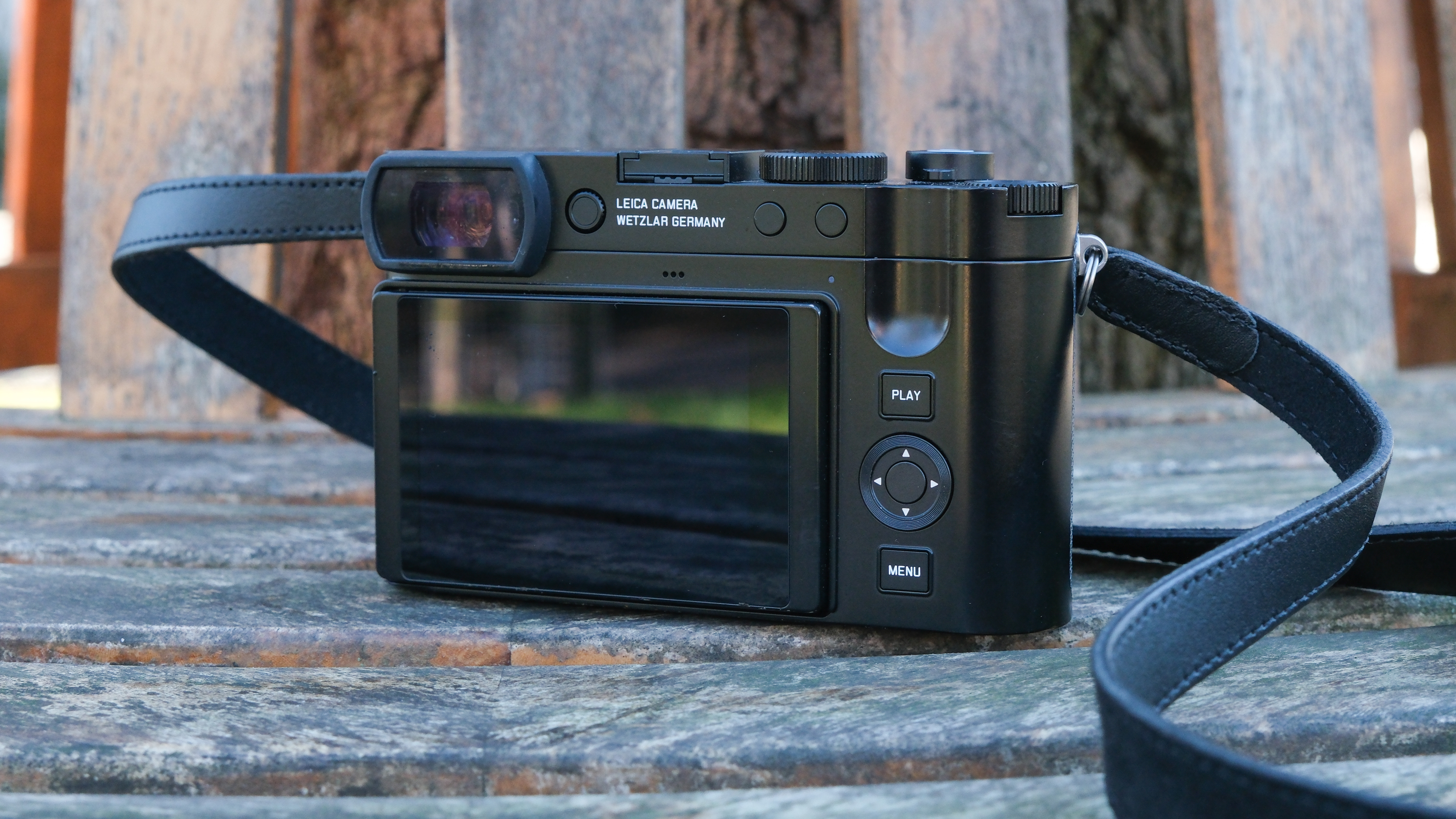 A Leica Q3 43 camera, with a strap attached, seen from behind showing the screen and buttons, sat on a wooden slatted bench