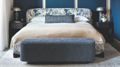 Blue bedroom with blue panelled walls, patterned white and blue bed, blue ottoman, and matching dark wood nightstands with matching lamps with metallic shades.