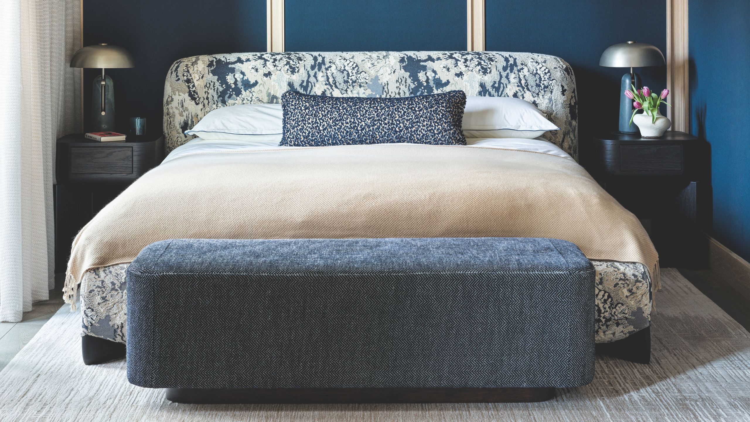 Blue bedroom with blue panelled walls, patterned white and blue bed, blue ottoman, and matching dark wood nightstands with matching lamps with metallic shades.