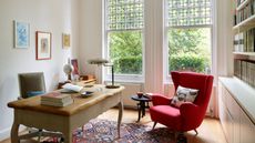 Bright white small home office with wooden table, bright white windows, art on the walls, comfortable red reading chair and built in storage on the right with shelves and cabinets