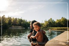 mother holding smiling infant while in swimwear next to a lake about to go swimming