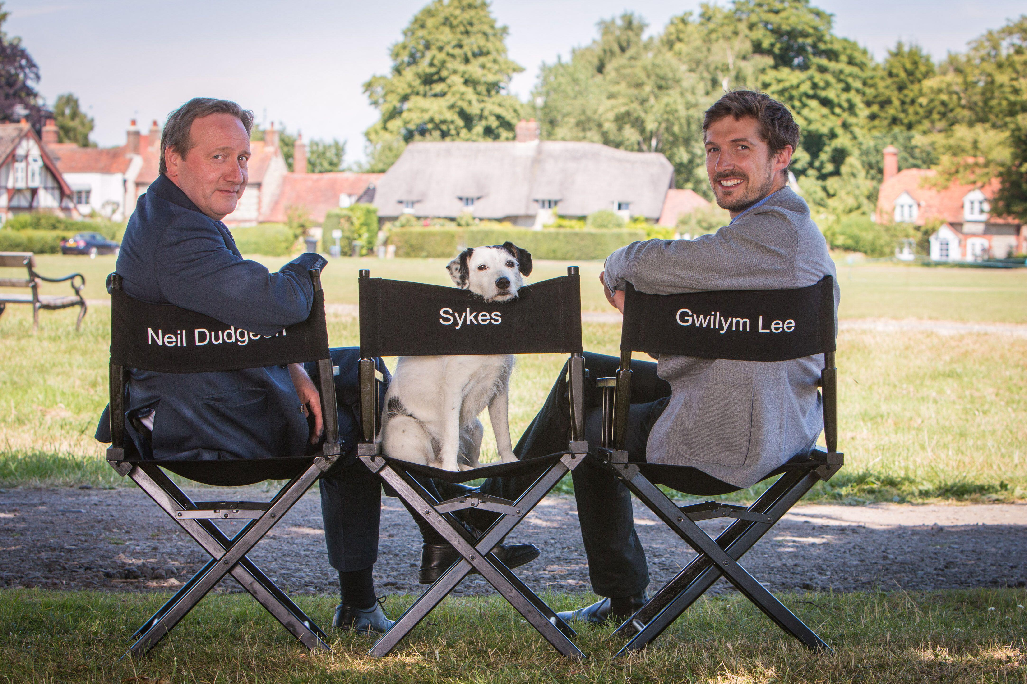 Neil Dudgeon and Gwilym Lee sit on black director's chairs on an outdoor film set with a black and white dog inbetween them