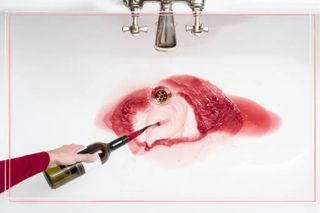 A bottle of red wine being poured down a sink