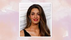 Image of Amal Clooney smiling wearing red lipstick and with curled long brunette hair at the front of one shoulder, wearing a black V-neck gown, on a pink and purple watercolour background
