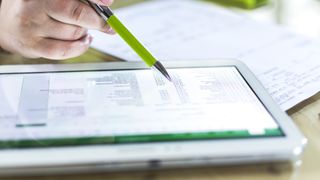 A hand holding a pen over a tablet, on which a spreadsheet is displayed.