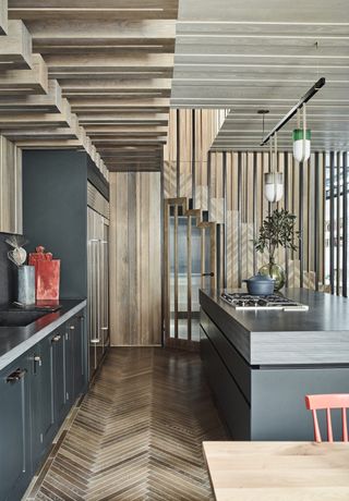 gray and wood kitchen with herringbone wood flooring, gray cabinetry, pendant lights, red chair, wooden ceiling, wooden panelling and floating wooden staircase