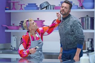 A contestant picks a crumb out of host Joel McHale's beard while standing in front of rows of pots and pans, in 'Crime Scene Kitchen.'