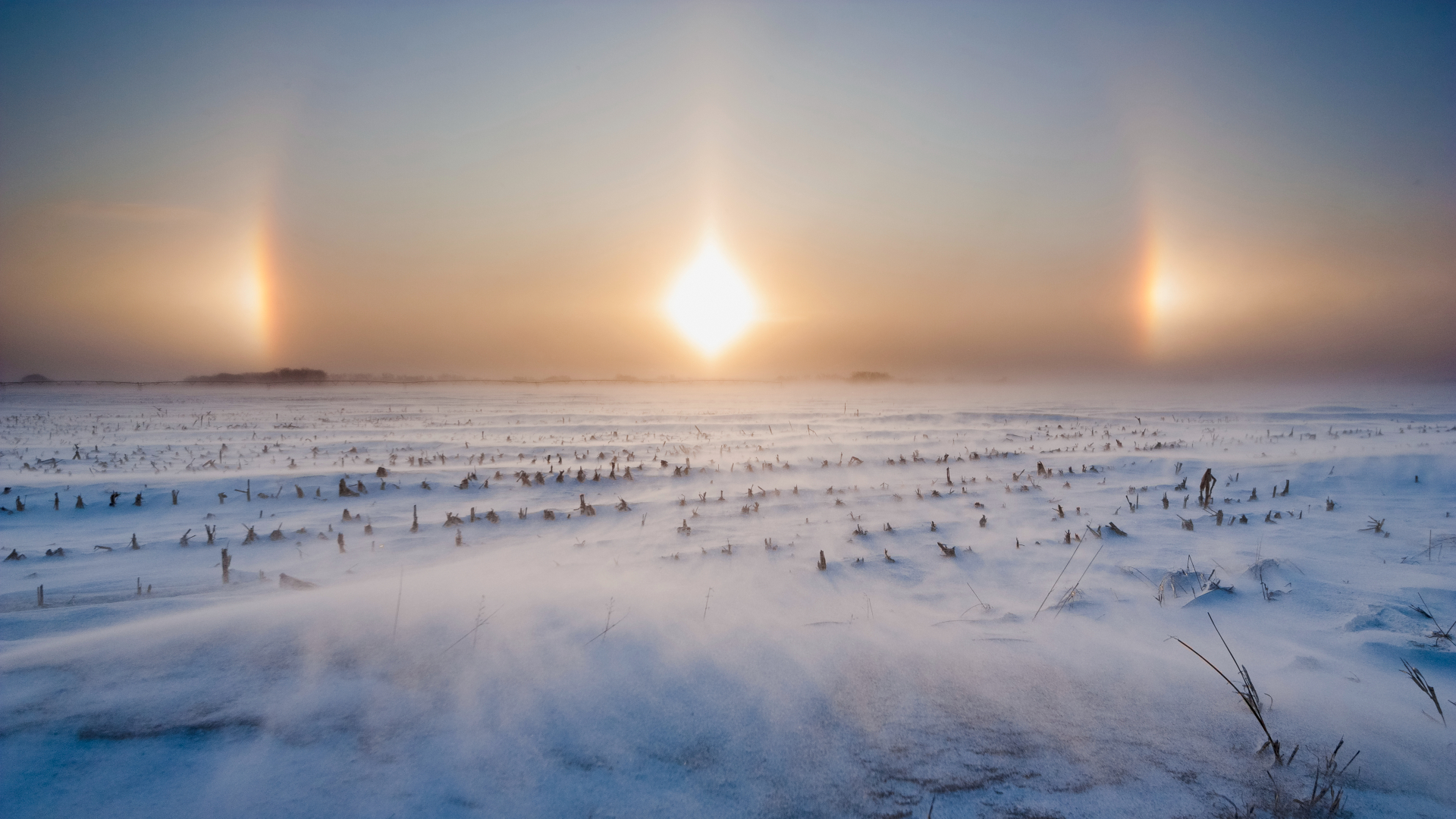 two bright pillars of light flank the sun on either side. The windswept landscape is covered in snow.