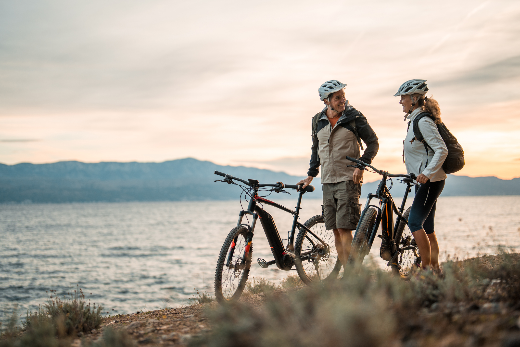 A mature Caucasian pair, wearing helmets and backpacks, stands beside their electric mountain bikes, conversing as the early sun illuminates the sea behind them.