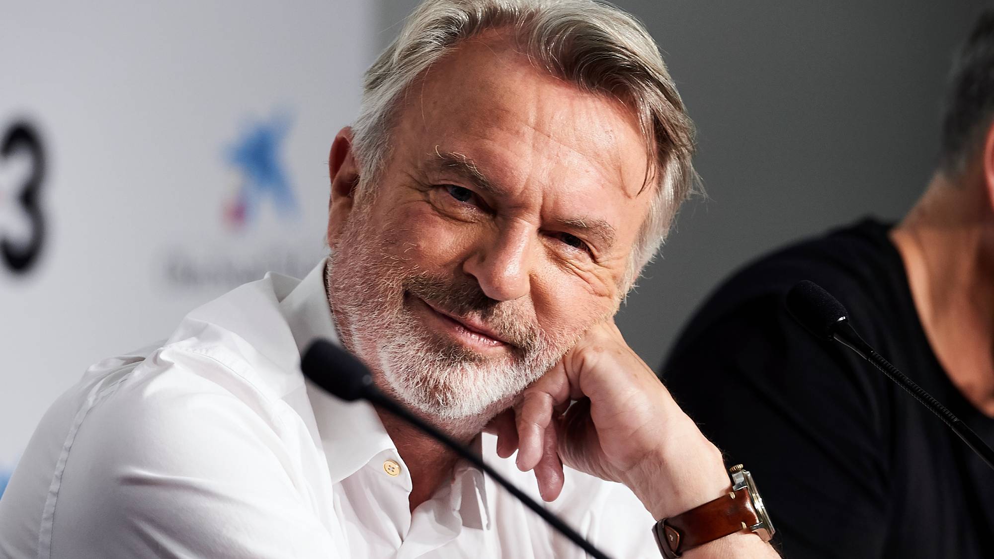 Sam Neill rests his head on his hand and looks toward the camera
