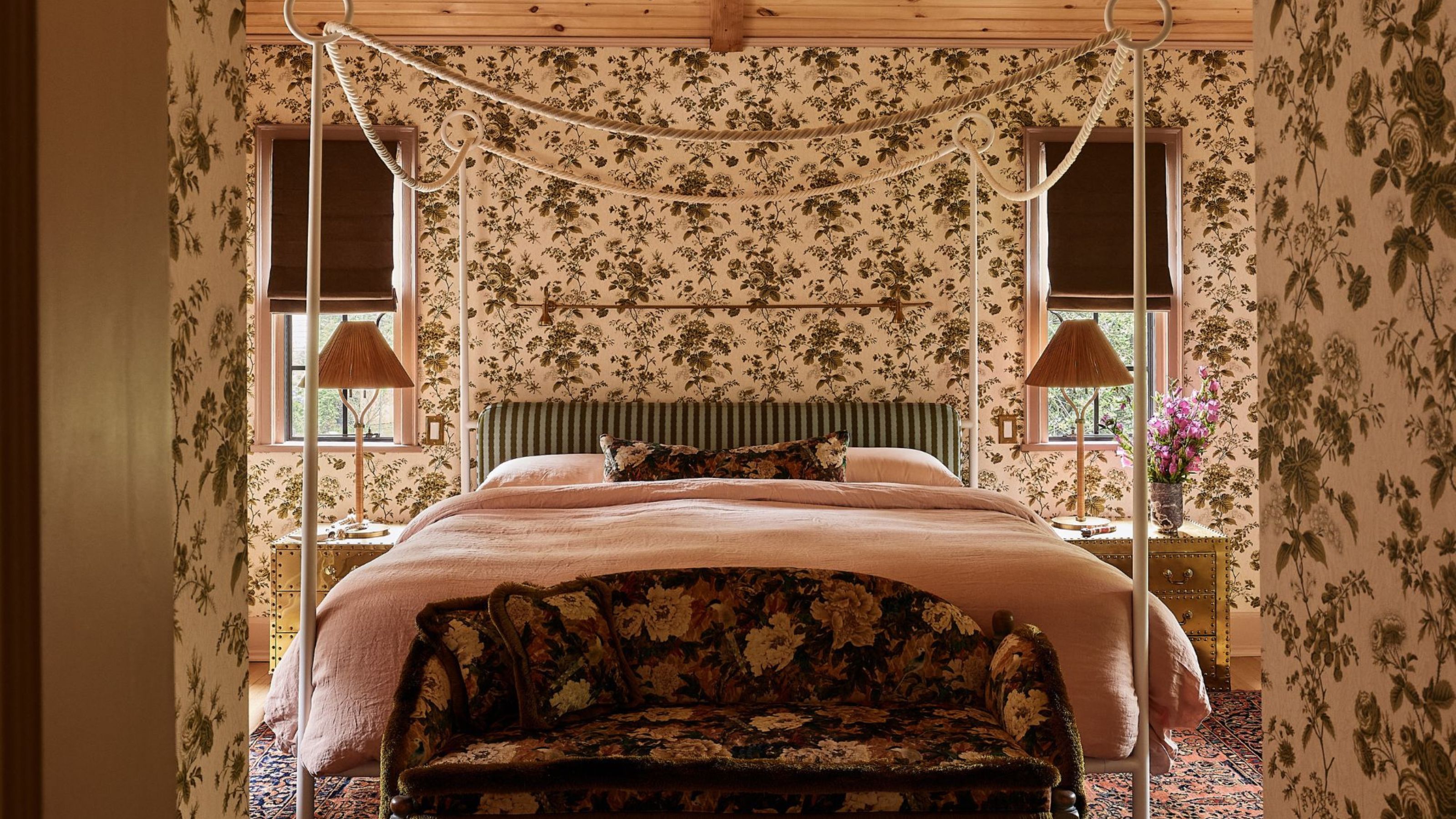a wallpapered primary bedroom with green/brown floral print walls, a pine clad ceiling and a four poster canopy bed with rope detailing 