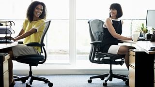 Two women chat while sitting in office chairs. 