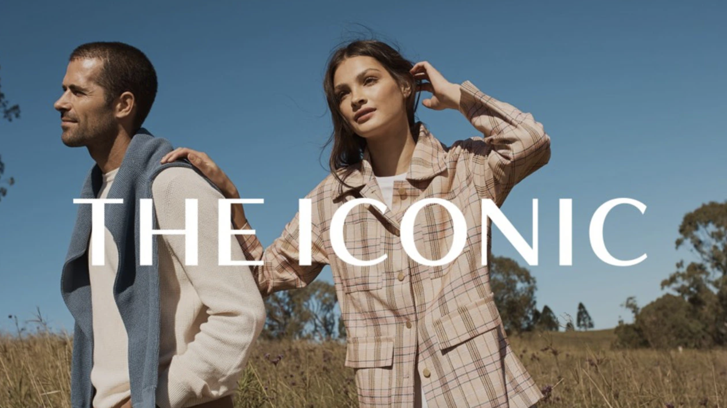 A man and a woman standing in a field with the women&#039;s hand on the man&#039;s shoulder with white writing that says The Iconic across the middle