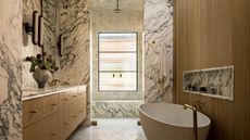 A large bathroom with warm wood finishes on the vanity and bath tub wall, and marble drenched across the walls and shower bench