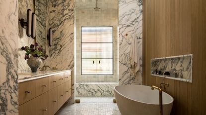 A large bathroom with warm wood finishes on the vanity and bath tub wall, and marble drenched across the walls and shower bench
