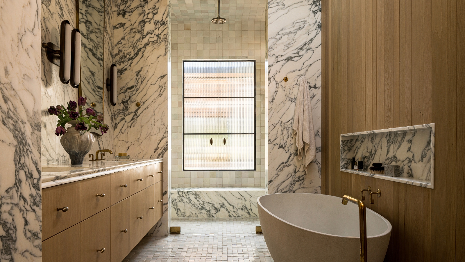 A large bathroom with warm wood finishes on the vanity and bath tub wall, and marble drenched across the walls and shower bench