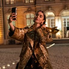 lily collins as emily taking a selfie and holding a pair of gold heels wearing a gold coat outside in europe at night in emily in paris season 6