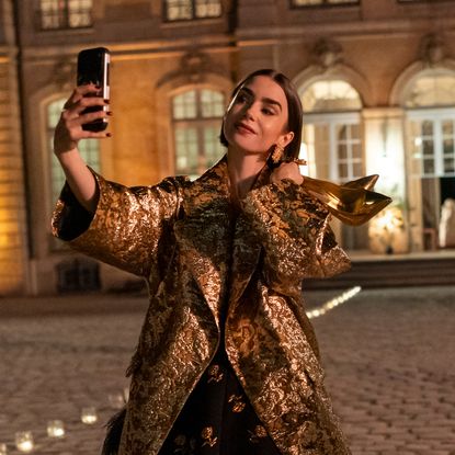 lily collins as emily taking a selfie and holding a pair of gold heels wearing a gold coat outside in europe at night in emily in paris season 6