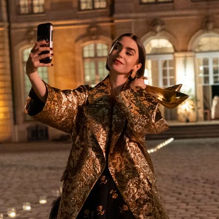 lily collins as emily taking a selfie and holding a pair of gold heels wearing a gold coat outside in europe at night in emily in paris season 6