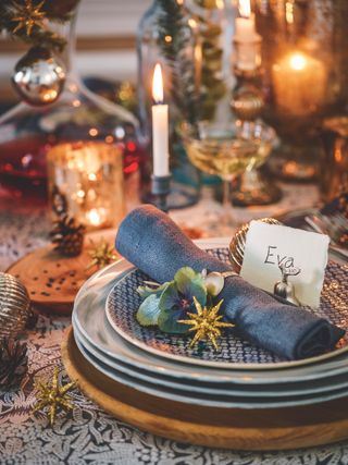 Close up of a place setting at a table for a Christmas meal with name card, decorations, drinking glasses and lit candles