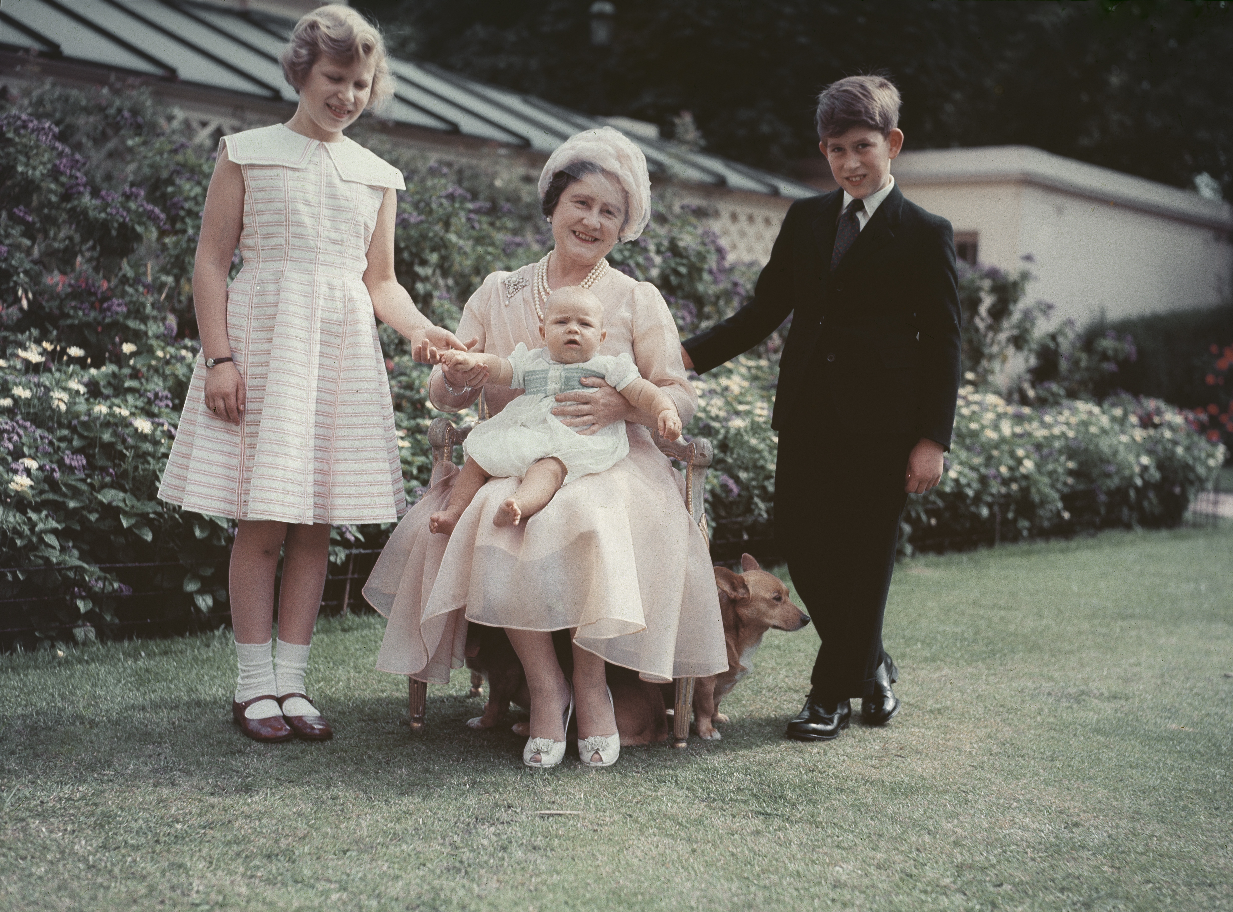 The Queen Mother sitting on a chair outside holding Prince Andrew, with Princess Anne and Prince Charles standing next to her in front of flowers