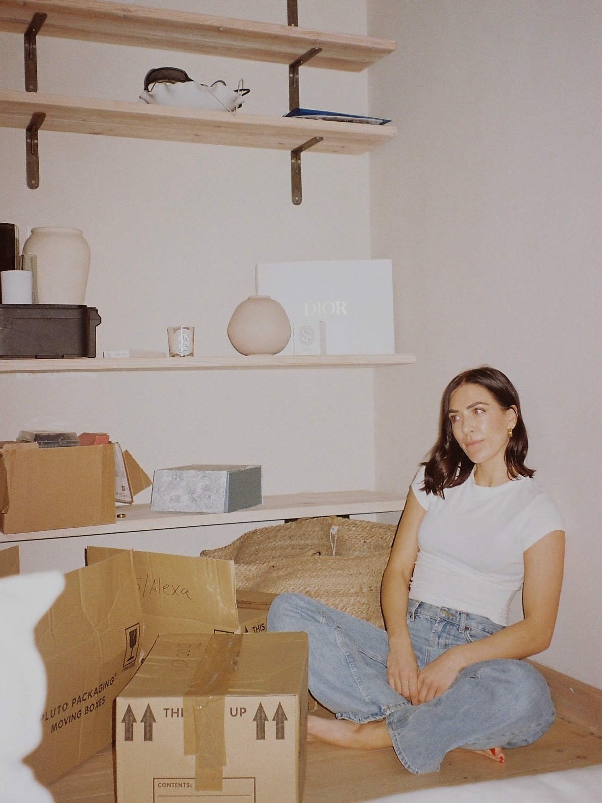 A woman sat on the floor of a living room with moving boxes, with a shiny chestnut brown bob