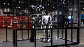 Sliver humanoid robot stands in a glass frame with a Tesla logo.