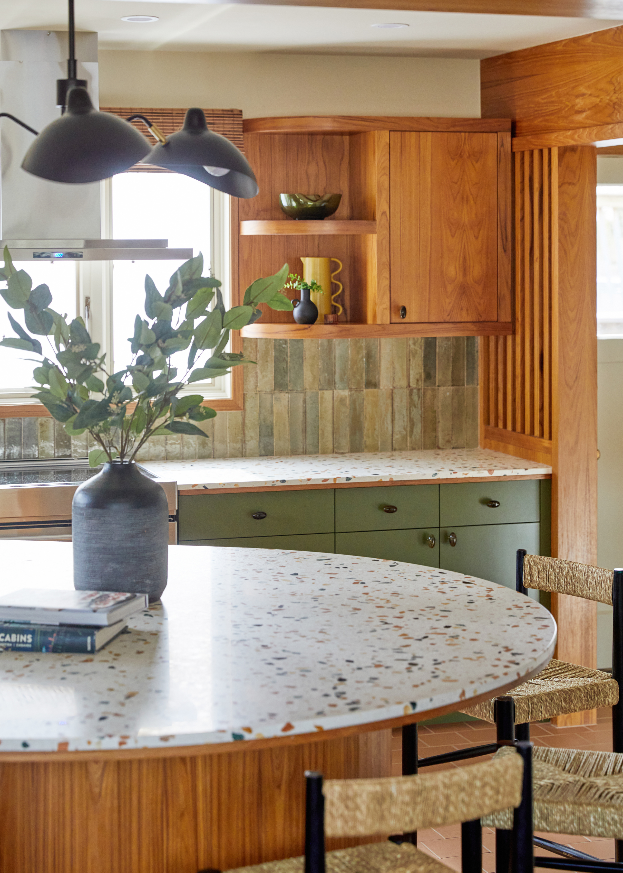 A bright kitchen with wood shelving and cabinets, a tile backsplash, a terrazzo countertop, and green cabinets, by a terrazzo top kitchen island with a vase of stems, a stack of books, and a row of woven stools