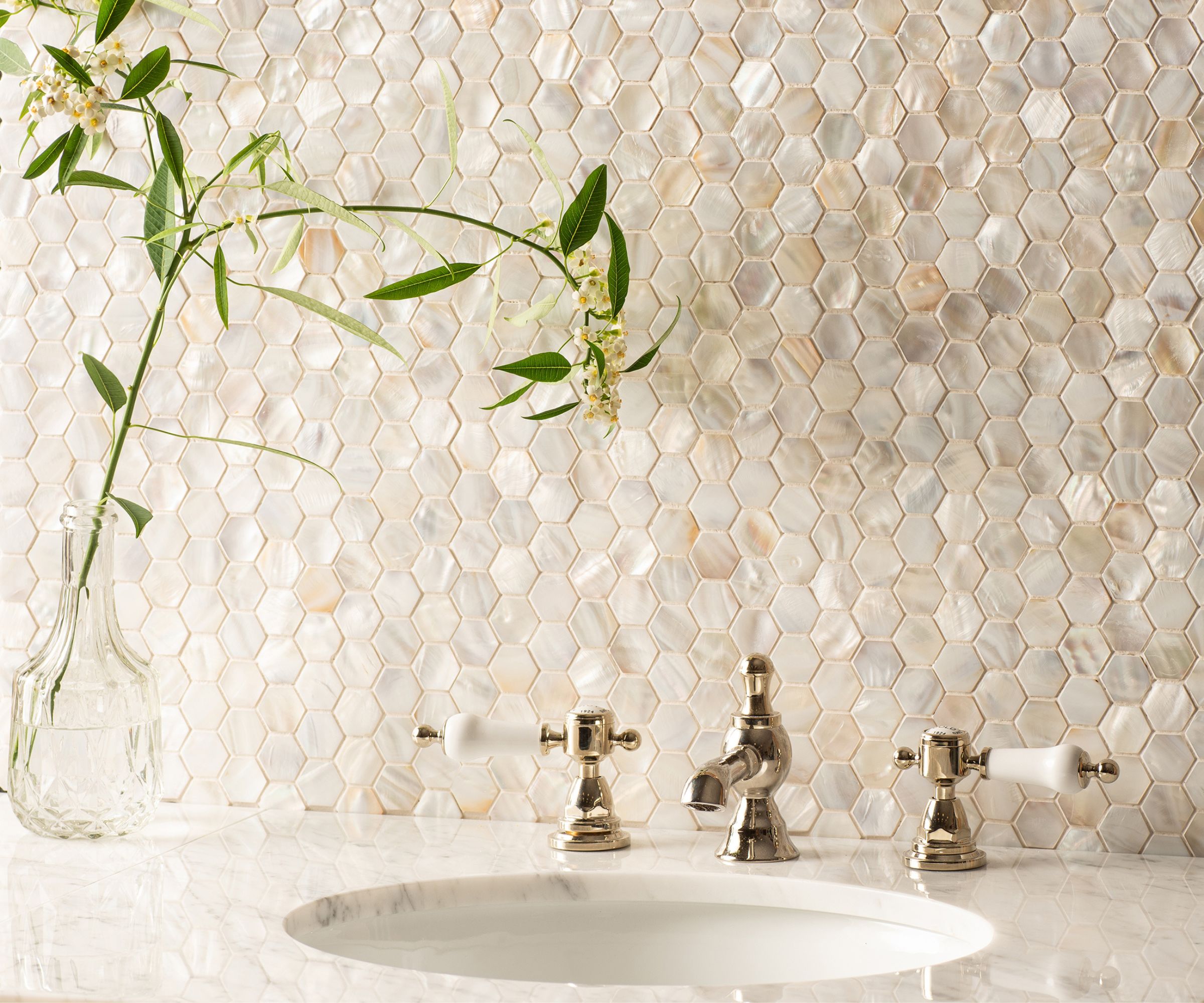 A close-up of a luxurious iridescent mother-of-pearl hexagonal mosaic tile wall behind a white marble basin with polished nickel three-piece basin taps. A glass vase with leafy flowering stems is positioned to the left on the marble surround.