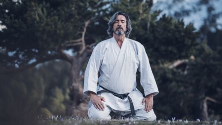An older man wearing a martial arts black belt kneels and looks very focused and relaxed.