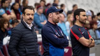 (L to R) Jason Sudeikis as Ted Lasso, Brendan Hunt as Coach Beard and Brett Goldstein as Roy Kent, standing on the sidelines, in Ted Lasso season 3