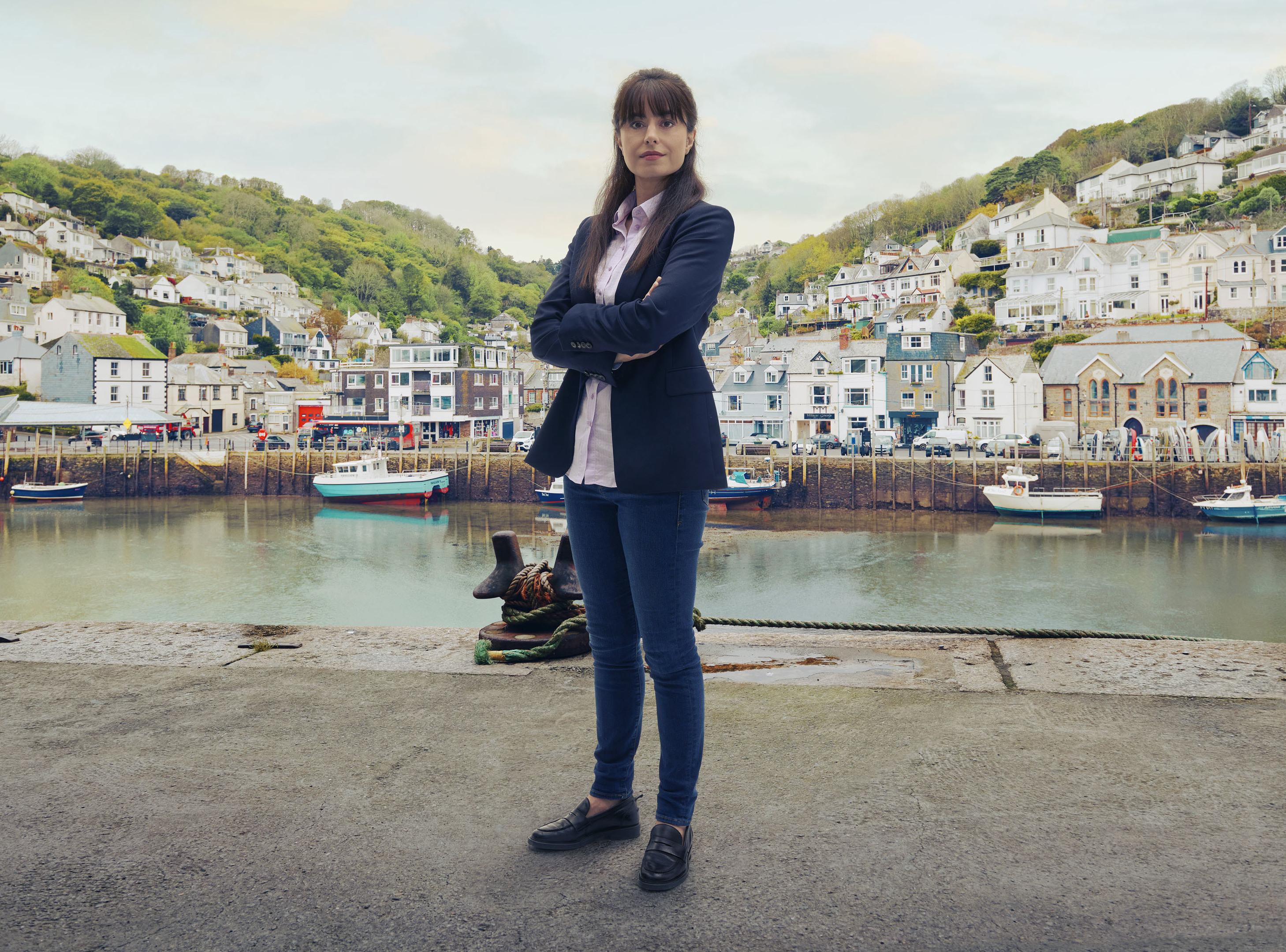 A shot of Zahra Ahmadi in character as DS Esther Williams, standing in the harbour of Shipton Abbott with the ocean and the houses going up the cliffs in the background behind her