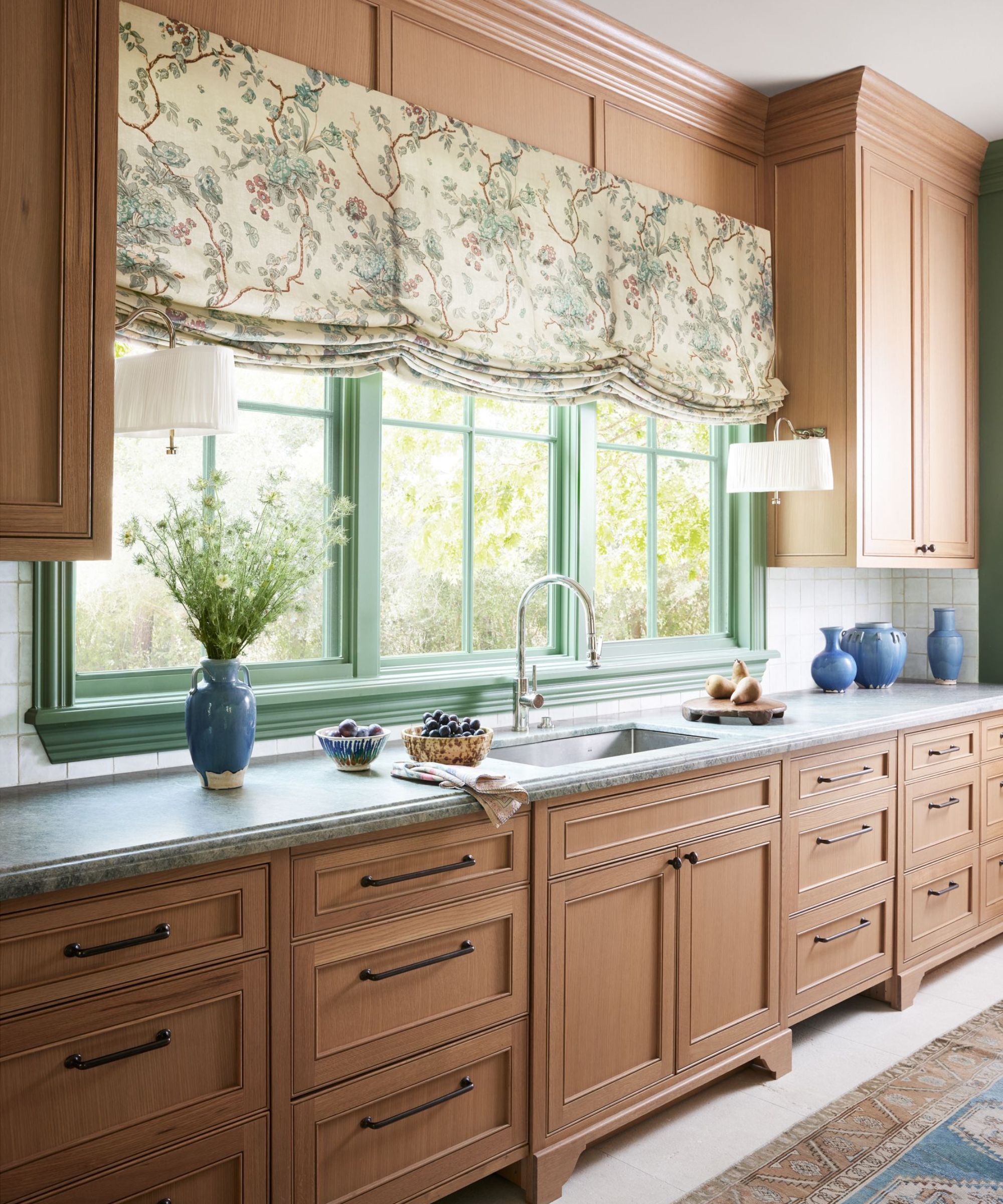 A kitchen with wooden cabinets, a triple window with green trim, and a floral roman blind in front of the sink.