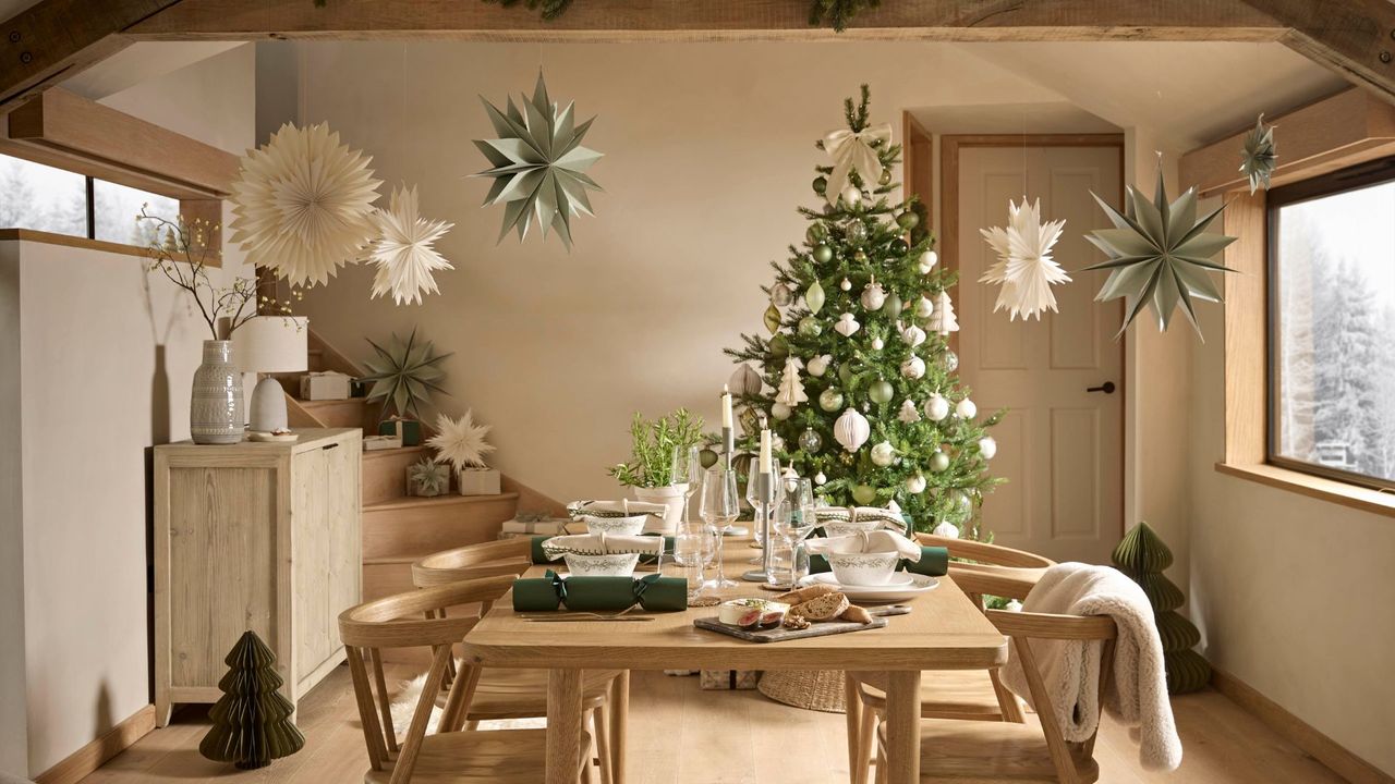 A minimalist dining room with Christmas stars hanging from the ceiling, a Christmas tree with neutral ornaments in the corner, and a wood dining set in the middle with paper Christmas crackers, cutlery and crockery
