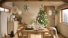 A minimalist dining room with Christmas stars hanging from the ceiling, a Christmas tree with neutral ornaments in the corner, and a wood dining set in the middle with paper Christmas crackers, cutlery and crockery