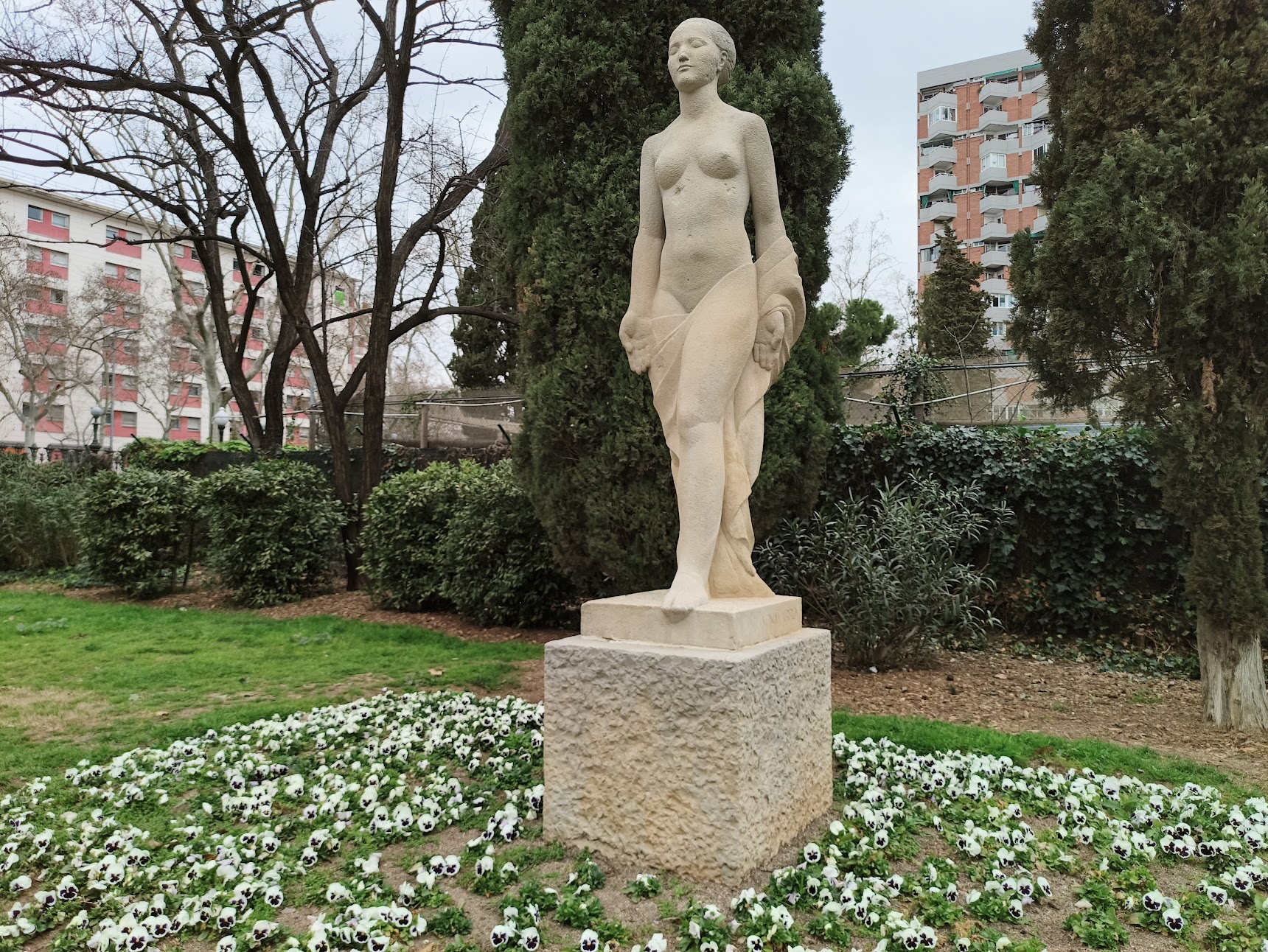 A stone statue of a serene, draped woman stands on a pedestal in a garden, surrounded by a bed of white flowers and tall evergreen trees.