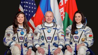 three smiling astronauts (two women and one man) in white spacesuits sit in front of their national flags against a dark background
