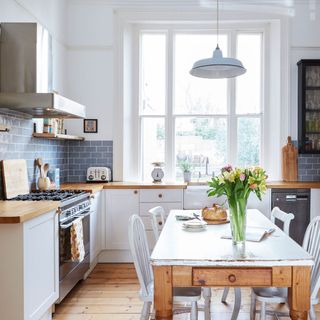 A kitchen with white cupboards and wooden tops, blue brick splashback, large window above a sink and a wooden dining table