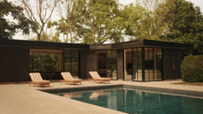 a small black bungalow pool house with large Crittal doors sits in front of a pool with three striped sunloungers
