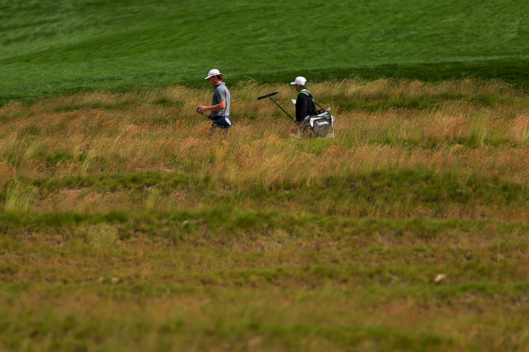 Noah Kent and his caddie walking through the long rough at Oakmont Country Club for the 2025 US Open, with only the top half of their bodies visible