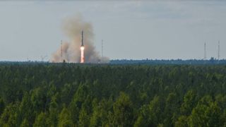 A russian soyuz rocket launches into a gray sky with green grass in the foreground