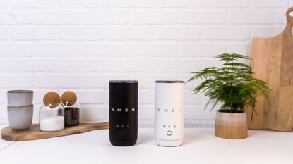 Image of a white and a black milk frother on a white kitchen countertop. There are two wooden cutting boards with espresso cups and plant vases on the counter.