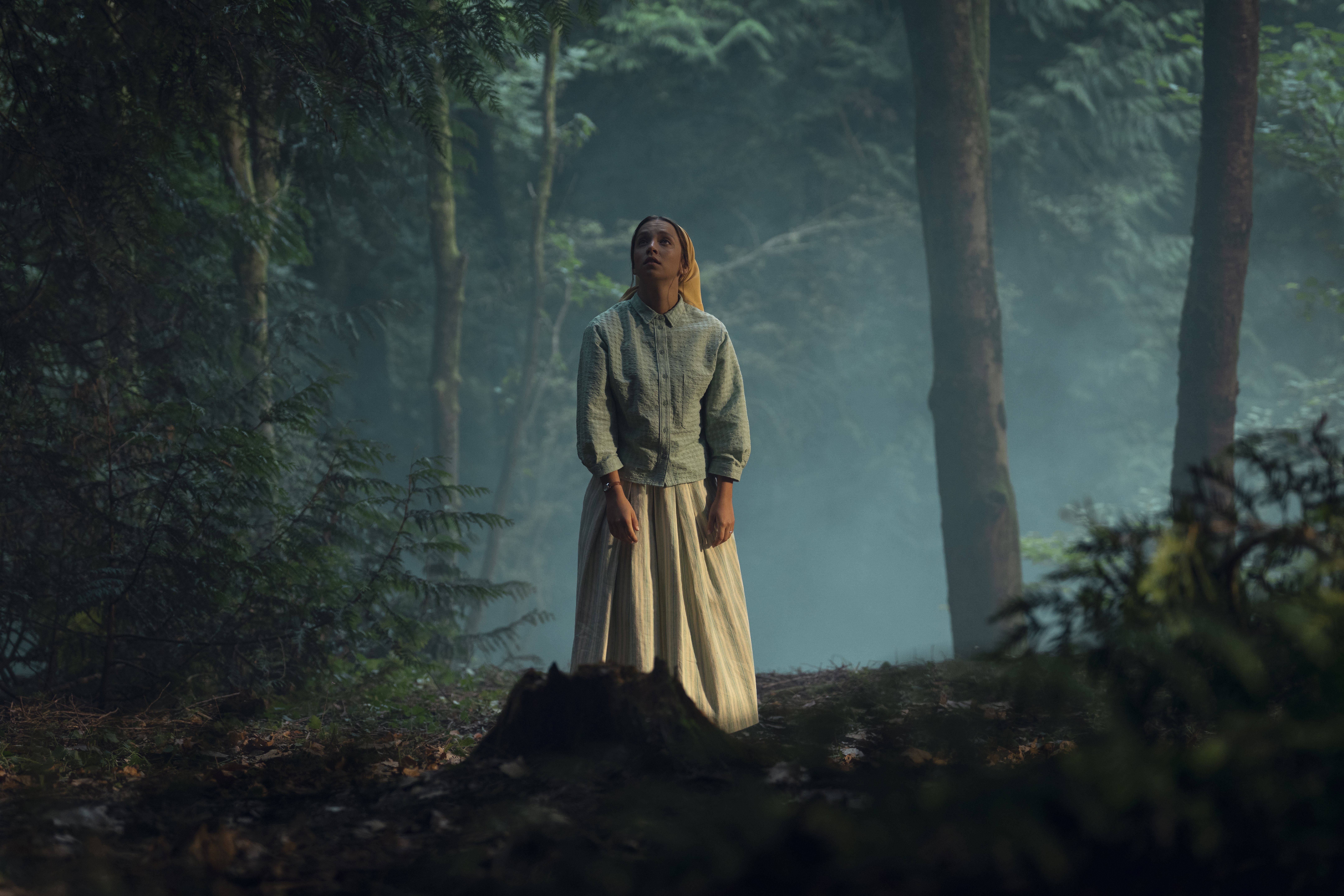 a woman in traditionalist clothing stands in a dark foggy wood looking upwards in a still from netflix's unchosen